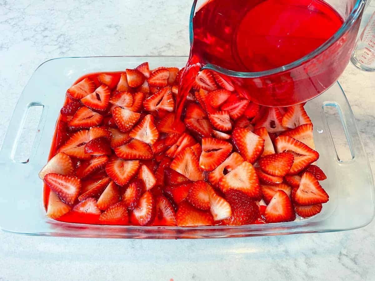 pouring strawberry jello over sliced strawberries layered into glass baking dish