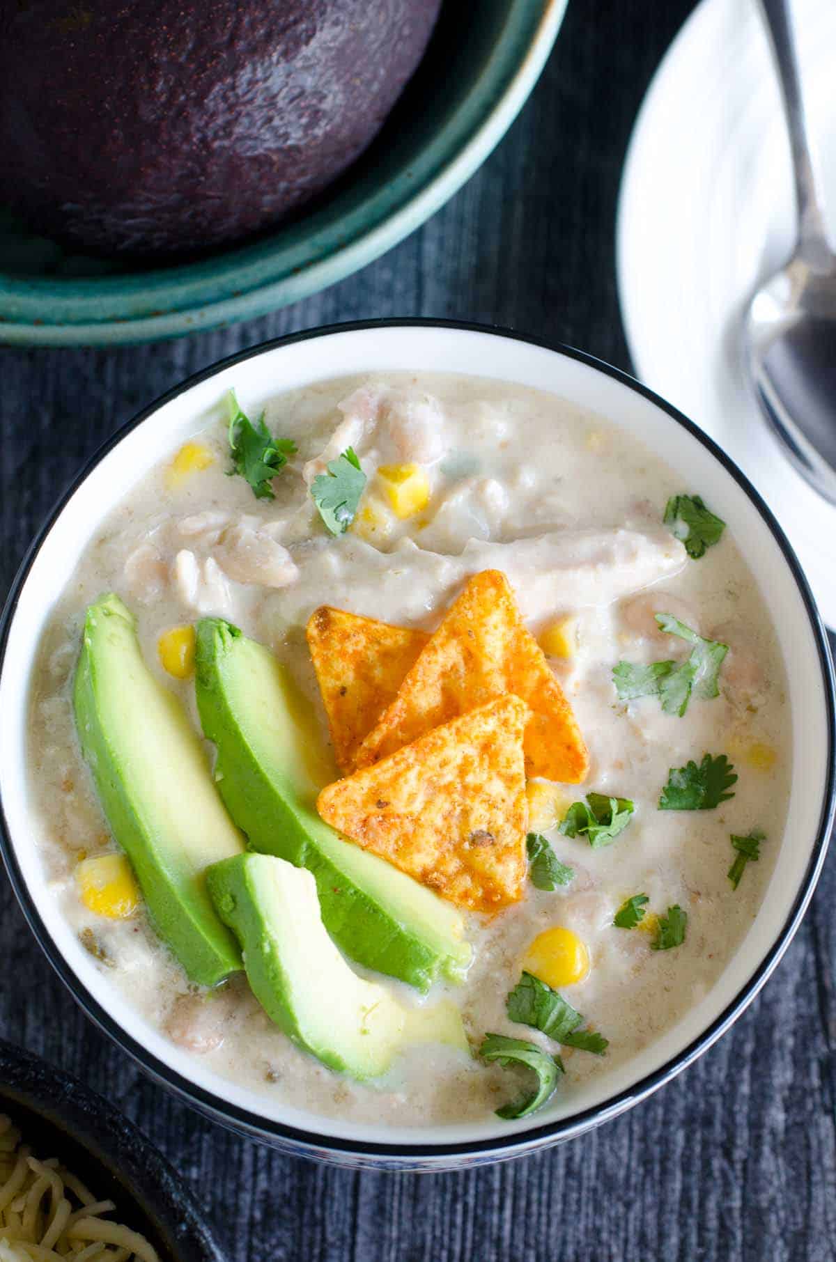bowl of white chicken chili topped with sliced avocados and chips